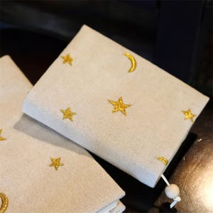 May include: Beige fabric pouch with gold embroidered stars and a crescent moon. The pouch has a wooden bead and string closure. The celestial design is repeated on the front of the pouch.
