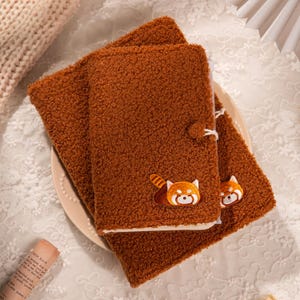 Pode incluir: Capa de caderno castanha e fofa com uma aplicação de panda vermelho. A capa tem um fecho de botão e é feita de um material macio e texturizado. O caderno está sobre um prato branco.