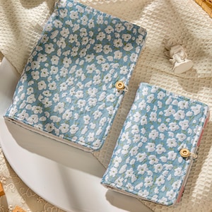 Pode incluir: Duas capas de livros com padrão floral. As capas são azul claro com um design floral branco e um fecho de botão dourado. As capas são de tamanhos diferentes, uma maior e outra menor, e provavelmente são para um diário ou caderno.