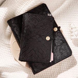 Pode incluir: Conjunto de diário e caderno preto com um padrão texturizado em forma de onda. O diário tem dois botões de madeira para fechar. O conjunto é exibido em um prato branco e uma superfície branca texturizada.