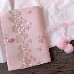 May include: A light pink journal with floral embroidery and a pearl clasp. The journal is decorated with embroidered flowers and leaves in shades of pink, white, and gold. A pair of pink pom-poms with a ribbon are also in the image.