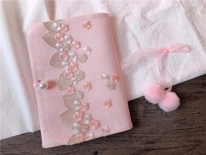 May include: A light pink journal with floral embroidery and a pearl clasp. The journal is decorated with embroidered flowers and leaves in shades of pink, white, and gold. A pair of pink pom-poms with a ribbon are also in the image.