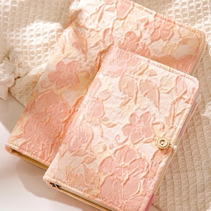 May include: Two floral patterned notebooks in shades of peach and cream. The covers feature a raised floral design and a button closure. The notebooks are likely for journaling or planning.