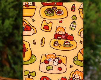 Puede incluir: Un cuaderno rectangular con una cubierta amarilla que presenta un patrón repetido de pandas rojos de dibujos animados en varias escenas. Las escenas incluyen comer, dormir y disfrutar de bocadillos. El cuaderno se muestra sobre una superficie de madera.