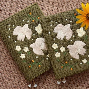 May include: Two olive green fabric covers featuring embroidered light beige birds, white flowers, and small orange berries. The textured material has a crinkled appearance, and each cover includes small dangling white flower-shaped charms at the bottom.