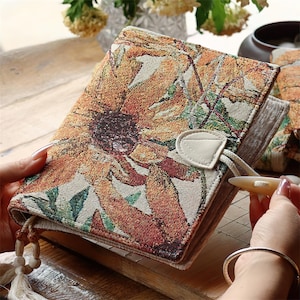 May include: A rectangular journal with a woven tapestry cover featuring a large sunflower pattern in shades of orange, yellow, green, and brown. The journal includes a cream-colored fabric closure with a toggle button and a decorative tassel with beads.