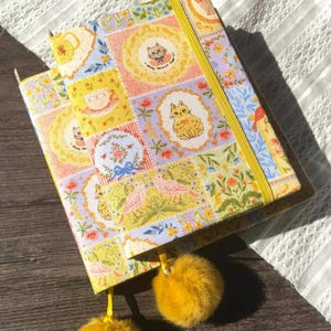 Puede incluir: Dos diarios decorativos con coloridos estampados de patchwork con gatos, flores y tazas de té. Los diarios tienen cierres elásticos amarillos y borlas de pompón amarillas a juego. Las cubiertas son una mezcla de colores pastel.