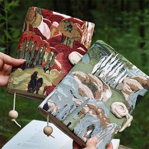 Può includere: Due copertine di libri decorative con disegni di arazzi paesaggistici. Una copertina presenta una scena desertica rossa, marrone e verde, mentre l'altra mostra una scena di montagna verde, blu e marrone. Ogni copertina ha un segnalibro con perline di legno e una chiusura con bottone in tessuto.
