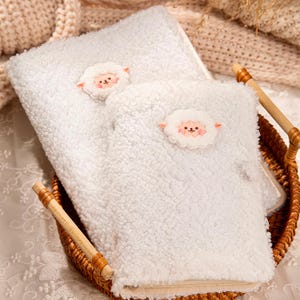 Pode incluir: Conjunto de toalha e luva de banho brancos e fofos, estilo sherpa. Cada peça apresenta um pequeno rosto de ovelha bordado. O conjunto é apresentado numa cesta de vime com alças de madeira, ideal para um spa ou casa de banho.