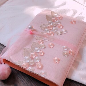 May include: A pink fabric case with floral embroidery and embellishments. The case features a pearl accent, a sheer ribbon, and two fluffy pink pom-poms. The design includes embroidered leaves and delicate flower appliqu&eacute;s.