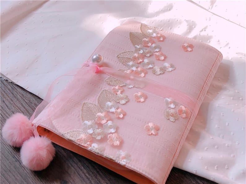 May include: A pink fabric case with floral embroidery and embellishments. The case features a pearl accent, a sheer ribbon, and two fluffy pink pom-poms. The design includes embroidered leaves and delicate flower appliqu&eacute;s.