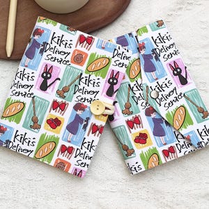 May include: A white fabric case with a colorful pattern featuring illustrations from "Kiki's Delivery Service." The design includes Kiki, Jiji the cat, brooms, bread, and bows. The case is secured with a button closure.
