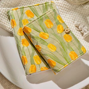 Pode incluir: Duas capas de livros com padrão floral com tulipas amarelas sobre fundo verde claro. As capas são fechadas com um botão de madeira. As capas estão sobre uma superfície branca.