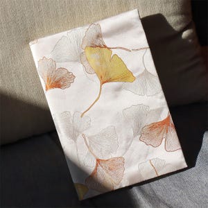 May include: Cream-colored fabric with a repeating pattern of stylized ginkgo leaves in shades of gold, orange, and gray. The leaves are detailed with delicate lines, creating a textured appearance. The fabric is folded, suggesting it could be a pillowcase or a similar textile item.