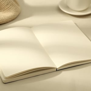 May include: An open notebook with blank, grid-lined pages. The pages are a soft cream color, and the notebook has a black spine. A cup and saucer are in the background. The notebook is ideal for journaling or note-taking.