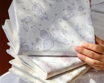 May include: A stack of notebooks with butterfly and constellation patterns on the covers. The covers are a light cream color with silver and lavender butterfly designs. The notebooks are stacked on top of each other.