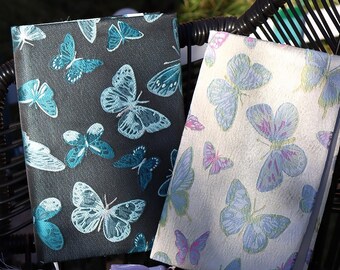 May include: Two fabric-covered notebooks with butterfly patterns. One is dark gray with teal butterflies, the other is off-white with blue and pink butterflies. Both have decorative tassels.