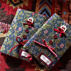 May include: Two rectangular fabric covers featuring a blue and green floral tapestry pattern. Each cover is decorated with three red buttons, a red velvet ribbon bow, and a white crocheted accent. A small white label reads "happy everyday".