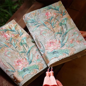 May include: Two rectangular journals covered in light green velvet fabric featuring intricate embroidery of pink lotus flowers, green leaves, and thin stems. The journals are accented with two soft pink tassels hanging from the bottom edge.
