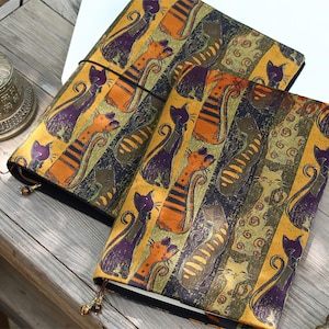 May include: Two rectangular fabric notebook covers featuring a repeating pattern of stylized cats in shades of orange, purple, and dark green. The covers are secured with thin black elastic bands and include small decorative beads on the bottom edges.