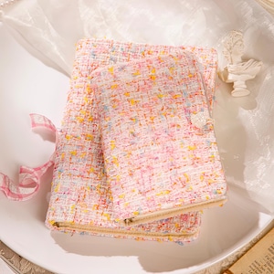 Puede incluir: Dos cuadernos de tweed rosa y blanco con cierre de cremallera y un botón en forma de corazón. Los cuadernos tienen un patrón texturizado colorido con hilos amarillos, azules y naranjas. Una cinta rosa está atada alrededor de uno de los cuadernos.