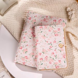 May include: A floral-patterned journal and matching fabric, featuring a delicate design of pink flowers on a white background. The journal has a button closure. The fabric is folded and placed next to the journal.