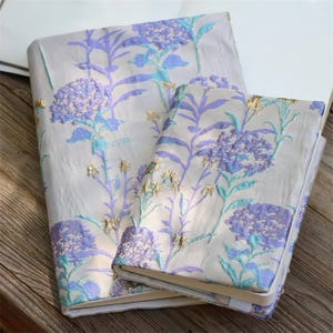 May include: Two notebooks with floral designs. The covers feature a light background with embroidered purple, blue, and gold floral patterns. The notebooks are rectangular and appear to be closed, with a ribbon bookmark.
