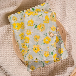 Pode incluir: Dois cadernos com padrão floral de flores amarelas e folhas verdes sobre um fundo azul claro. Os cadernos estão fechados e presos com um botão de madeira e uma corda. Eles estão em um prato bege.