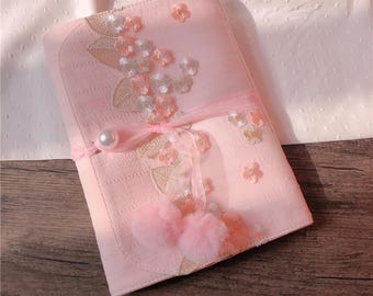May include: A light pink, rectangular journal or notebook with floral embroidery and a ribbon closure. The cover features embroidered flowers in white, peach, and gold, with two small pink pom-poms and a pearl accent.