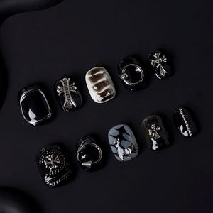 Puede incluir: Un conjunto de diez uñas postizas negras y plateadas con varios diseños, incluyendo cruces, anillos y tachuelas.