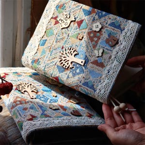 May include: A fabric-covered journal featuring a geometric pattern in blue, tan, and red. The cover is adorned with wooden cutouts of a tree, a bird, a butterfly, and flowers, finished with white lace trim and two wooden beads on a cord.