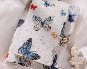 May include: A white journal and matching notepad set with a butterfly print in shades of blue and red. The journal has a wooden button closure. The set is on a white surface.