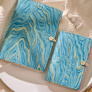 May include: Two blue journals with a gold marbled design. The journals are closed with a button and loop closure. The journals are on a white plate.