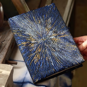 Könnte beinhalten: Ein rechteckiges, marineblaues Notizbuch mit strukturiertem Einband. Der Einband zeigt ein radiales Design in Weiß, Gold und Blau, das einem Lichtausbruch ähnelt. Das Notizbuch wird von einer Hand gehalten.