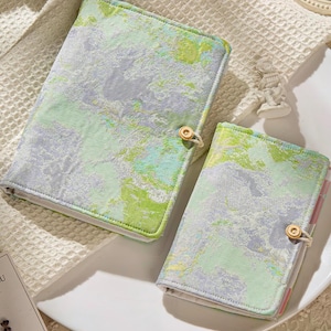 Pode incluir: Dois cadernos revestidos de tecido com um design semelhante a aguarela em tons de verde, cinza e azul. Os cadernos têm um fecho de botão e uma encadernação de cor clara. O caderno maior tem cerca de 20 cm de altura e o menor cerca de 15 cm.