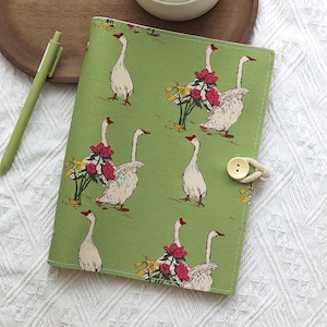 May include: A green journal with a pattern of white geese with red beaks and accents of red flowers. The journal has a button closure with a beige rope. A green pen is visible in the background.