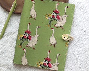 May include: A green journal with a pattern of white geese with red beaks and accents of red flowers. The journal has a button closure with a beige rope. A green pen is visible in the background.