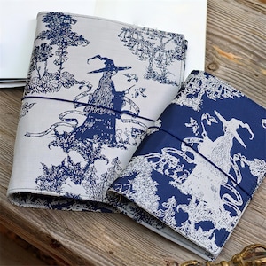 May include: Two fabric-covered journals featuring a blue and white witch design. Each journal is secured with a blue elastic band. The witch design is set against a backdrop of trees and foliage. The journals are on a wooden surface.