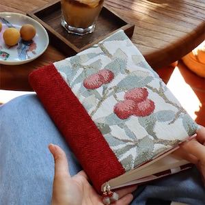 May include: A patterned book cover with a red spine and a fabric design of red berries and green leaves on a white background. The book is open, showing the pages.