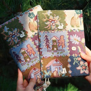 May include: Two rectangular fabric notebook covers featuring a woven tapestry pattern of houses, trees, flowers, and fruit. The covers are decorated with small wooden buttons shaped like a rabbit, a cat, and an apple, with dangling wooden beads.