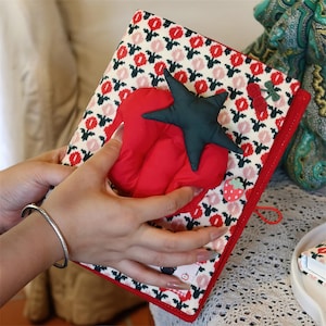 May include: A fabric-covered notebook featuring a repeating pattern of red and black flowers on a cream background. The cover is adorned with a plush red tomato-shaped accent topped with a dark green star-shaped stem, a small cherry charm, and a strawberry patch.