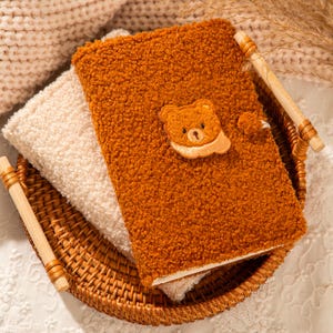 Puede incluir: Un diario marrón y esponjoso con una aplicación de oso de peluche. El diario descansa sobre una cesta tejida con un libro color crema debajo. El diario tiene una cubierta de felpa texturizada y un cierre de madera.