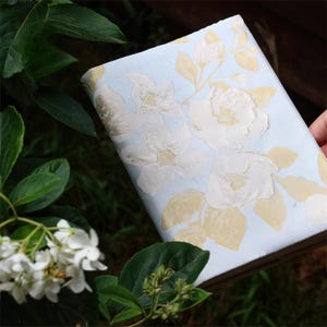 May include: A roll of light blue floral wallpaper with large white and pale yellow flowers. The wallpaper has a textured, embossed design. The flowers and leaves are arranged across the surface. The wallpaper is partially unrolled.