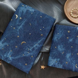 Puede incluir: Dos cuadernos azules cubiertos de tela con un diseño celestial. Las cubiertas presentan estrellas doradas y lunas crecientes dispersas sobre un fondo nublado, similar al denim. Los cuadernos están colocados sobre una superficie gris plateado brillante.
