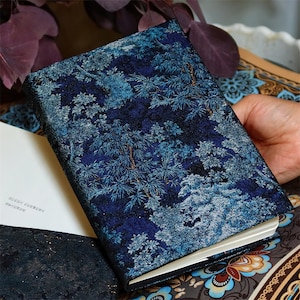 Puede incluir: Un diario de tapa dura con una cubierta azul oscuro que presenta un patrón detallado y texturizado de árboles y follaje estilizados en tonos azules y plateados. El diario abierto revela páginas blancas. La estética general es elegante e inspirada en la naturaleza.