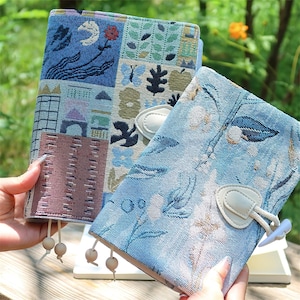 May include: Two decorative notebooks with patterned covers. One notebook features a patchwork design with blue, green, and brown panels. The other notebook has a blue floral pattern. Both notebooks have a white closure and wooden bead accents.