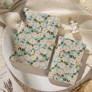 May include: Two floral-patterned book covers. The covers feature a light blue background with white flowers and green leaves. Each cover has a wooden button closure. The covers are placed on a white plate.