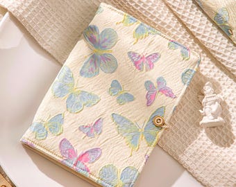 May include: A cream-colored notebook cover with a butterfly pattern in shades of blue, pink, and yellow. The cover has a button closure and is made of textured fabric. The notebook is placed on a white surface.