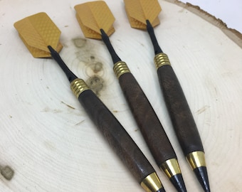 May include: Four darts with dark brown wooden handles, brass accents, and black tips. The dart flights are a mustard yellow color. The darts are arranged on a light-colored wooden surface.