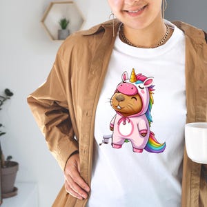 以下が含まれることがあります： ユニコーンのコスチュームを着た漫画のビーバーが描かれた白いTシャツ。ビーバーはピンクのワンピース、金色の角、虹色のたてがみと尾を身に着けています。デザインは気まぐれでカラフルです。
